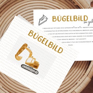 Könnte beinhalten: Zwei Karten mit dem deutschen Wort "BÜGELBILD" in Gold gedruckt. Eine Karte zeigt eine Aquarellillustration eines gelben Baggers. Die andere Karte enthält Anweisungen und ein Bügeleisensymbol.