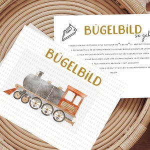 Könnte beinhalten: Zwei Karten mit Text und einer Aquarellillustration einer Lokomotive. Eine Karte trägt das Wort "BÜGELBILD" in goldfarbenen Buchstaben. Die andere Karte enthält Anweisungen und die Worte "BÜGELBILD so geht's". Die Karten liegen auf einem geflochtenen Korb.