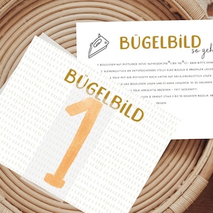 Könnte beinhalten: Eine weiße Karte mit dem Text "Bügelbild so geht's" in goldenen Buchstaben. Die Karte enthält Anweisungen zur Verwendung eines Bügelbildes. Die Karte liegt auf einem geflochtenen Korb mit weißem Hintergrund.