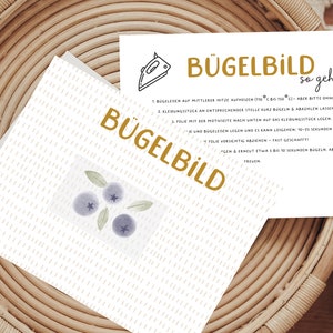 Könnte beinhalten: Eine weiße Karte mit dem Wort "BÜGELBILD" in goldfarbenen Buchstaben, mit einem Blaubeer-Design. Eine weitere Karte mit Anweisungen und den Worten "BÜGELBILD so geht's" ist ebenfalls sichtbar. Die Karten liegen auf einem geflochtenen Korb.