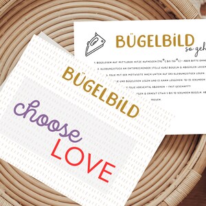 Könnte beinhalten: Eine weiße Karte mit dem Text "BÜGELBILD choose LOVE" in lila und roter Schrift. Die Karte liegt auf einem geflochtenen Korb mit einer kleineren weißen Karte mit Anweisungen zum Aufbügeln eines Designs. Die Anweisungen sind in Deutsch und beinhalten den Text "BÜGELBILD so geht's".