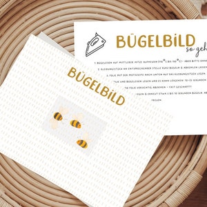 Könnte beinhalten: Eine weiße Karte mit dem Text "Bügelbild so geht's" in goldenen Buchstaben. Die Karte enthält Anweisungen zum Aufbügeln eines Bildes. Auf der Karte sind drei gelbe und schwarze Bienen abgebildet.