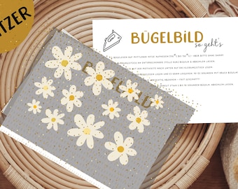 Può includere: Un cartoncino grigio con sfondo bianco e glitter dorati. Il cartoncino presenta un motivo di margherite bianche e il testo "Bügelbild so geht's" in lettere dorate. Il testo sotto il titolo spiega come utilizzare un trasferimento da stirare.