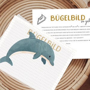 Könnte beinhalten: Eine weiße Karte mit Anweisungen zur Verwendung eines Bügelbildes. Die Karte hat eine goldene Grafik eines Bügeleisens und den Text "Bügelbild so geht's". Die Karte liegt in einem geflochtenen Korb mit einer Illustration eines blauen und weißen Delfins.