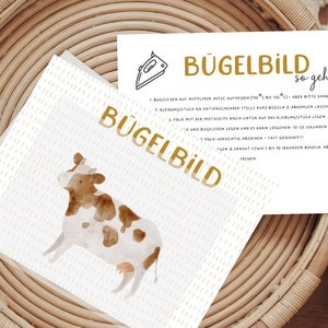 Könnte beinhalten: Eine weiße Karte mit goldener Schrift, die "Bügelbild so geht's" lautet und Anweisungen zum Aufbügeln eines Bügelbildes enthält. Die Karte liegt auf einem geflochtenen Korb mit einer Illustration einer braunen und weißen Kuh.