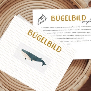 Könnte beinhalten: Eine weiße Karte mit einer Illustration eines blauen Wals und dem Text "BÜGELBILD" in goldenen Buchstaben. Die Karte enthält auch Anweisungen zum Aufbügeln eines Bügelbildes, die in deutscher Sprache verfasst sind.
