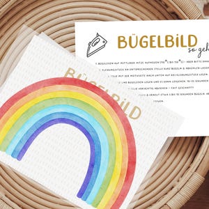 Könnte beinhalten: Anleitung zum Aufbügeln eines Bügelbildes mit einem Regenbogen-Design. Die Anleitung ist in deutscher Sprache verfasst und enthält Schritte wie "Bügelbild mit der bedruckten Seite nach unten auf den Stoff legen" und "10-20 Sekunden mit festem Druck bügeln".