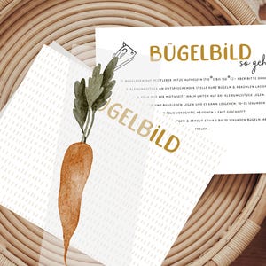 Könnte beinhalten: Eine Aquarellillustration einer Karotte mit grünen Blättern auf einer weißen Karte mit goldenem Text "BÜGELBILD so geht's". Die Karte liegt auf einem geflochtenen Korb.