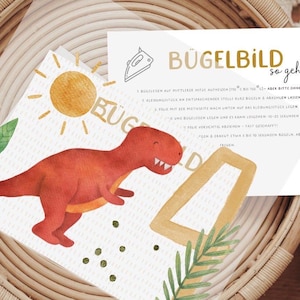 Könnte beinhalten: Eine weiße Karte mit dem Text "Bügelbild so geht's" und Anweisungen zur Verwendung von Bügelbildern. Die Karte liegt in einem geflochtenen Korb mit einer Dinosaurier- und einer Sonnenillustration.
