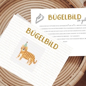 Könnte beinhalten: Eine weiße Karte mit dem Text "Bügelbild so geht's" in schwarzer und goldener Schrift. Die Karte enthält Anweisungen zum Aufbügeln eines Aufklebers. Die Karte liegt auf einem geflochtenen Korb mit einer weißen Karte mit dem Text "Bügelbild" in goldener Schrift und einem Comic-Tiger, der einen Partyhut trägt.