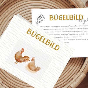 Könnte beinhalten: Eine weiße Karte mit dem Text "Bügelbild" in goldenen Buchstaben und Anweisungen, wie man ein Design aufbügelt. Die Karte liegt auf einem geflochtenen Korb mit einer Illustration eines braunen und weißen Hahns und einer Henne.