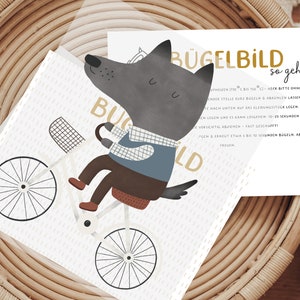 Könnte beinhalten: Eine weiße Karte mit einem grauen Wolf, der Fahrrad fährt. Der Wolf trägt ein blau-weiß kariertes Hemd und eine braune Hose. Die Karte hat den Text "BÜGELBILD so geht's" in goldenen Buchstaben. Die Karte liegt auf einem geflochtenen Korb.