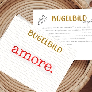 Könnte beinhalten: Eine weiße Karte mit dem Text "BÜGELBILD so geht's" in goldener Schrift. Die Karte enthält Anweisungen zum Aufbügeln eines Designs. Die Karte enthält auch den Text "BÜGELBILD amore." in goldener und roter Schrift.