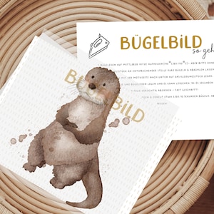 Könnte beinhalten: Eine weiße Karte mit Anweisungen zum Aufbügeln eines Bügelbildes. Die Karte trägt den Text "Bügelbild so geht's" in goldenen Buchstaben. Die Karte liegt auf einem geflochtenen Korb mit einer Illustration einer braunen und weißen Otter.