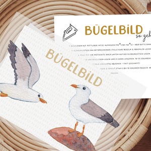 Könnte beinhalten: Aquarell-Illustration von zwei Möwen auf weißem Hintergrund, mit dem deutschen Text "BÜGELBILD" und Anweisungen. Das Bild befindet sich auf einem geflochtenen Korb. Die Möwen sind weiß, grau und orange.