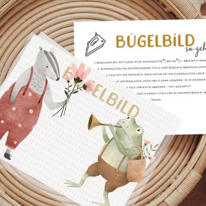 Könnte beinhalten: Eine weiße Karte mit Anweisungen zum Aufbügeln eines Bügelbildes. Die Karte zeigt einen Comic-Dachs, der einen Strauß rosa Blumen hält, und einen Comic-Frosch, der Trompete spielt. Der Text auf der Karte lautet "Bügelbild so geht's".