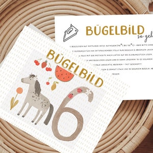Könnte beinhalten: Eine weiße Karte mit einer bunten Illustration eines Pferdes, einer Wassermelone, Kirschen und der Zahl 6. Die Karte trägt den Text "Bügelbild" in goldenen Buchstaben. Die Karte liegt auf einem geflochtenen Korb.