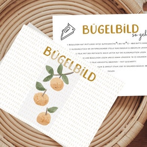 Könnte beinhalten: Eine weiße Karte mit dem Text "Bügelbild so geht's" in goldenen Buchstaben. Die Karte enthält Anweisungen zum Aufbügeln eines Bildes. Die Karte liegt auf einem geflochtenen Korb mit einer weißen Karte mit einer Zitronenbaumillustration.