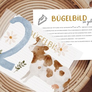 Könnte beinhalten: Ein Aquarelldesign mit einer blauen Zahl 2, einer Kuh und Gänseblümchen. Das Design enthält den Text "BÜGELBILD so geht's" und Bügelanweisungen. Das Bild ist vor einem gewebten Hintergrund platziert.