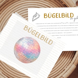 Könnte beinhalten: Zwei weiße Karten mit goldenem Text und einem Aquarell-Diskokugel-Design. Eine Karte trägt die Aufschrift "BÜGELBILD" und die andere enthält Anweisungen auf Deutsch. Die Karten liegen auf einem Weidenkorb.