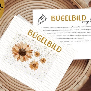 Könnte beinhalten: Eine weiße Karte mit dem Text "Bügelbild so geht's" und Anweisungen zum Aufbügeln eines Bügelbildes. Die Karte ist mit einem goldenen Glitzerhintergrund und vier kleinen Sonnenblumenillustrationen verziert.