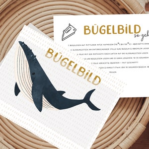 Könnte beinhalten: Eine weiße Karte mit einer Walillustration und dem Text "BÜGELBILD" in goldenen Buchstaben. Die Karte enthält auch Anweisungen zum Aufbügeln des Bildes, mit dem Text "so geht's" in schwarzen Buchstaben.