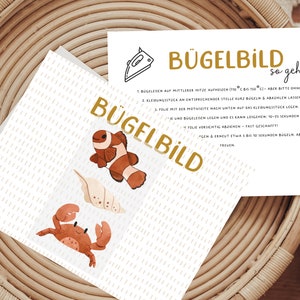 Könnte beinhalten: Eine weiße Karte mit dem Text "BÜGELBILD" und Bügelanweisungen. Die Karte zeigt Illustrationen eines Clownfisches, Muscheln und einer Krabbe. Der Hintergrund ist hellbeige mit einem geflochtenen Korb.