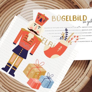 Könnte beinhalten: Ein farbenfroher Bügelbild mit einem Nussknacker, der eine Geschenkbox hält. Das Bügelbild enthält auch einen roten Strumpf mit einem weißen Pfeil und einem Blumenmuster. Die Anleitung für die Verwendung des Bügelbildes ist auf der Karte enthalten. "Bügelbild so geht's" steht in goldenen Buchstaben oben auf der Karte.