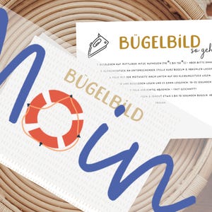 Könnte beinhalten: Ein weißer Bügelbild-Transferbogen mit einem rot-weißen Rettungsring-Motiv und dem Schriftzug BÜGELBILD MOIN in Gold und Blau. Eine beiliegende weiße Anleitungskarte zeigt ein Bügeleisen-Symbol und den Text BÜGELBILD so geht's mit nummerierten Schritten.