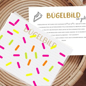 Könnte beinhalten: Eine weiße Karte mit bunten Streuseln und dem Wort "BÜGELBILD" in Gold. Eine zweite Karte mit Anweisungen und einer Bügeleisen-Grafik ist ebenfalls sichtbar. Die Karten liegen auf einer gewebten Oberfläche.