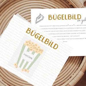 Könnte beinhalten: Eine weiße Karte mit dem Wort "BÜGELBILD" in goldfarbenen Buchstaben und einer Popcorn-Grafik. Die Karte liegt auf einem Weidenkorb. Eine zweite Karte mit Anweisungen ist ebenfalls sichtbar. Die Popcorn-Grafik ist ein grün-weiß gestreifter Behälter.