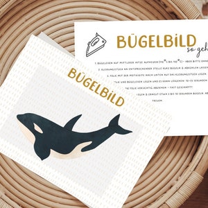 Könnte beinhalten: Eine weiße Karte mit dem Text "Bügelbild so geht's" in goldenen Buchstaben. Die Karte enthält Anweisungen zum Aufbügeln eines Bildes. Das Bild zeigt einen schwarz-weißen Orca.