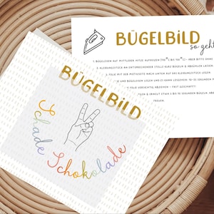 Könnte beinhalten: Zwei weiße Karten mit goldenem und regenbogenfarbenem Text und Illustrationen. Eine Karte sagt "BÜGELBILD" und "Schade Schokolade" mit einer Hand, die ein Friedenszeichen macht. Die andere Karte hat Anweisungen und die Worte "BÜGELBILD so geht's".
