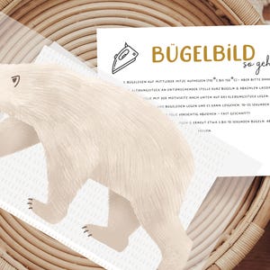 Könnte beinhalten: Eine weiße Karte mit schwarzem Text und einer goldenen Grafik eines Bügeleisens. Der Text lautet "Bügelbild so geht's" und enthält Anweisungen zum Bügeln eines Bügelbildes.