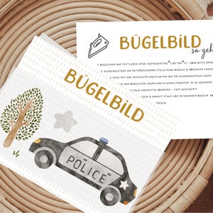 Könnte beinhalten: Eine weiße Karte mit den Worten "Bügelbild so geht's" in goldener Schrift. Die Karte enthält Anweisungen, wie man einen Aufkleber aufbügelt. Die Karte liegt auf einem geflochtenen Korb mit einem schwarz-weißen Polizeiauto-Aufkleber darauf.