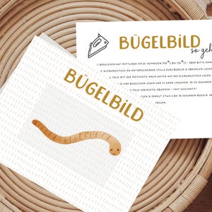 Könnte beinhalten: Anleitung zum Aufbügeln eines Bügelbildes auf Stoff. Die Anleitung ist in deutscher Sprache verfasst und enthält eine Comic-Illustration eines Bügeleisens und eines Wurms.