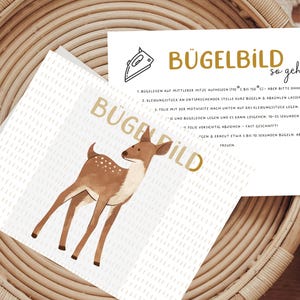 Könnte beinhalten: Eine weiße Karte mit einer braunen Hirsch-Illustration und dem Text "BÜGELDILD" in goldenen Buchstaben. Die Karte enthält auch Anweisungen zum Aufbügeln eines Bügelbildes.