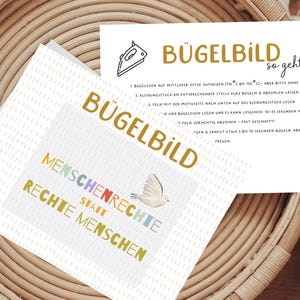 Könnte beinhalten: Zwei Karten mit Text und Illustrationen werden auf einer gewebten Oberfläche angezeigt. Eine Karte lautet "BÜGELBILD" und "MENSCHENRECHTE STATT RECHTE MENSCHEN" mit einer Taubengrafik. Die andere Karte enthält Anweisungen und die Worte "BÜGELBILD so geht's".