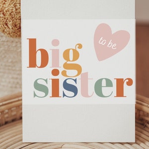 Könnte beinhalten: Eine weiße Karte mit dem Text "big sister" in bunten Buchstaben. Ein rosa Herz mit dem Text "to be" befindet sich über dem Text.