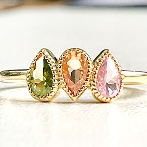 May include: A gold ring featuring three pear-shaped gemstones. The gemstones are green, peach, and pink.