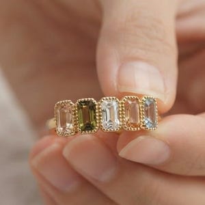 May include: A gold ring featuring five rectangular, emerald-cut gemstones in a row. The stones display a gradient of colors including light peach, olive green, clear white, soft pink, and pale blue, all set within a beaded gold metal frame.
