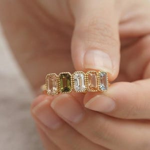 May include: A gold ring featuring five rectangular, emerald-cut gemstones arranged in a row. The stones display a gradient of colors including light peach, olive green, clear white, soft pink, and pale blue, all set within a beaded gold frame.