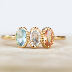 May include: A gold ring featuring three oval-shaped gemstones. The gemstones are light blue, white, and peach in colour.