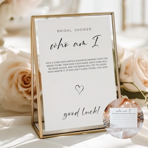 May include: A gold-framed sign for a bridal shower with the text "who am I" in elegant script. The sign includes instructions for a game, a heart symbol, and the words "good luck!" A smaller card with lines for writing is also visible.