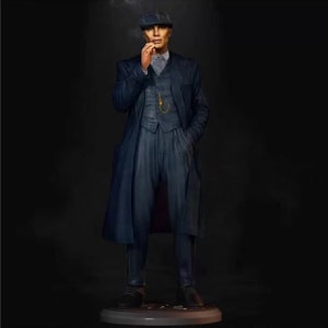 Op de afbeelding: Een blauw pak met een vest en een lange jas. De man draagt een platte pet en houdt een sigaret vast.