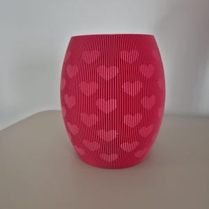 May include: A bright pink, cylindrical vase featuring a heart pattern. The vase is constructed from vertical lines, giving it a textured appearance. The heart design is repeated across the surface, adding a romantic touch to the decor.
