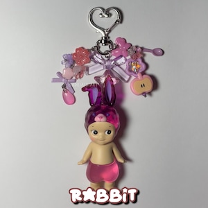 Puede incluir: Un llavero decorativo con una peque&ntilde;a mu&ntilde;eca que lleva un gorro de conejito morado y pantalones cortos rosas. La mu&ntilde;eca est&aacute; sujeta a un cierre plateado en forma de coraz&oacute;n con abalorios rosas y morados. La palabra "RABBIT" est&aacute; escrita en letras rosas en la parte inferior.