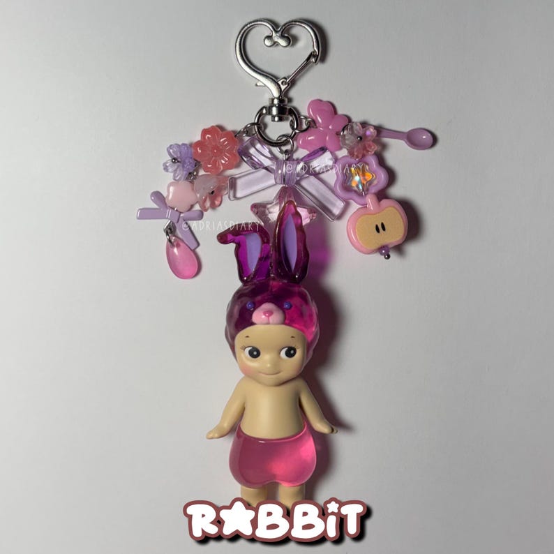 Puede incluir: Un llavero decorativo con una peque&ntilde;a mu&ntilde;eca que lleva un gorro de conejito morado y pantalones cortos rosas. La mu&ntilde;eca est&aacute; sujeta a un cierre plateado en forma de coraz&oacute;n con abalorios rosas y morados. La palabra "RABBIT" est&aacute; escrita en letras rosas en la parte inferior.