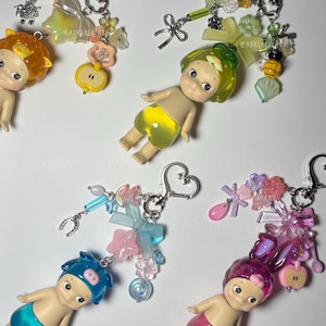Puede incluir: Una colecci&oacute;n de llaveros coloridos con peque&ntilde;as figuras de mu&ntilde;ecas y varios accesorios. Los llaveros tienen una variedad de dijes, incluyendo lazos, flores y cuentas. Las mu&ntilde;ecas son de diferentes colores, como amarillo, rosa y azul.