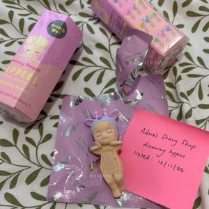 K&ouml;nnte beinhalten: Eine Sammlung von Sonny Angel Dreamy Hippers Figuren, darunter eine Figur im Giraffen-Design, Verpackungen und ein pinkfarbener Haftzettel mit handschriftlichem Text. Die Schachteln und Verpackungen sind haupts&auml;chlich rosa und lila.